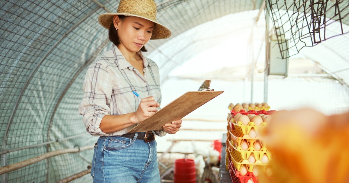 A woman writing on a clipboard near trays of eggs inside a poultry farm with chickens in the background.